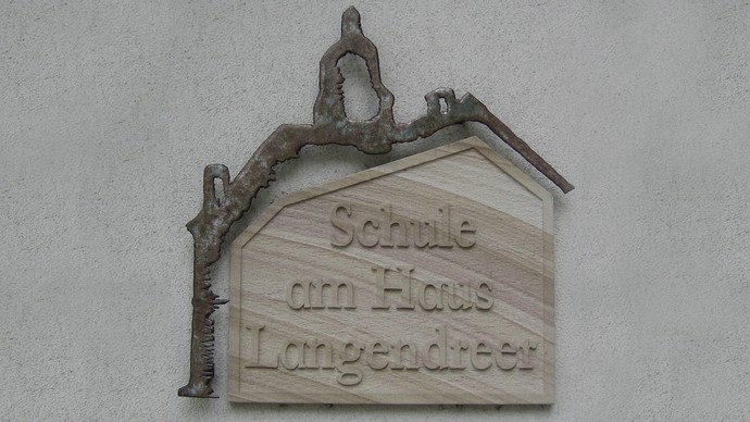 Das Foto zeigt eine Sandsteintafel mit der Aufschrift "Schule am Haus Langendreer". Über der Sandsteintafel befindet sich eine Metall-Nachbildung der Silhouette des Glockenturms von Haus Langendreer. Das Foto zeigt eine Sandsteintafel mit der Aufschrift "Schule am Haus Langendreer". Über der Sandsteintafel befindet sich eine Metall-Nachbildung der Silhouette des Glockenturms von Haus Langendreer.