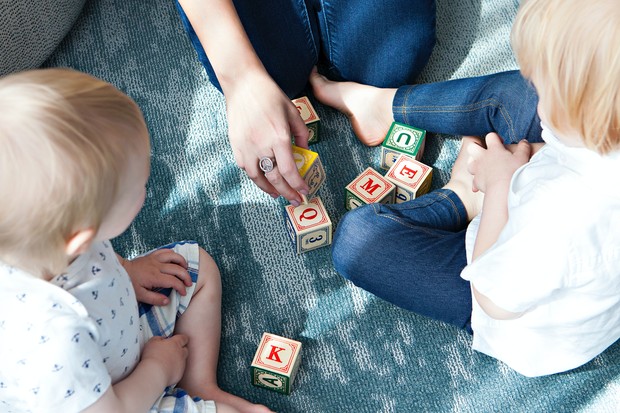 Zwei Kleinkinder sitzen zusammen mit einer erwachsenen Person am Boden und spielen mit mit Buchstaben bedruckten Würfeln. Zwei Kleinkinder sitzen zusammen mit einer erwachsenen Person am Boden und spielen mit mit Buchstaben bedruckten Würfeln.